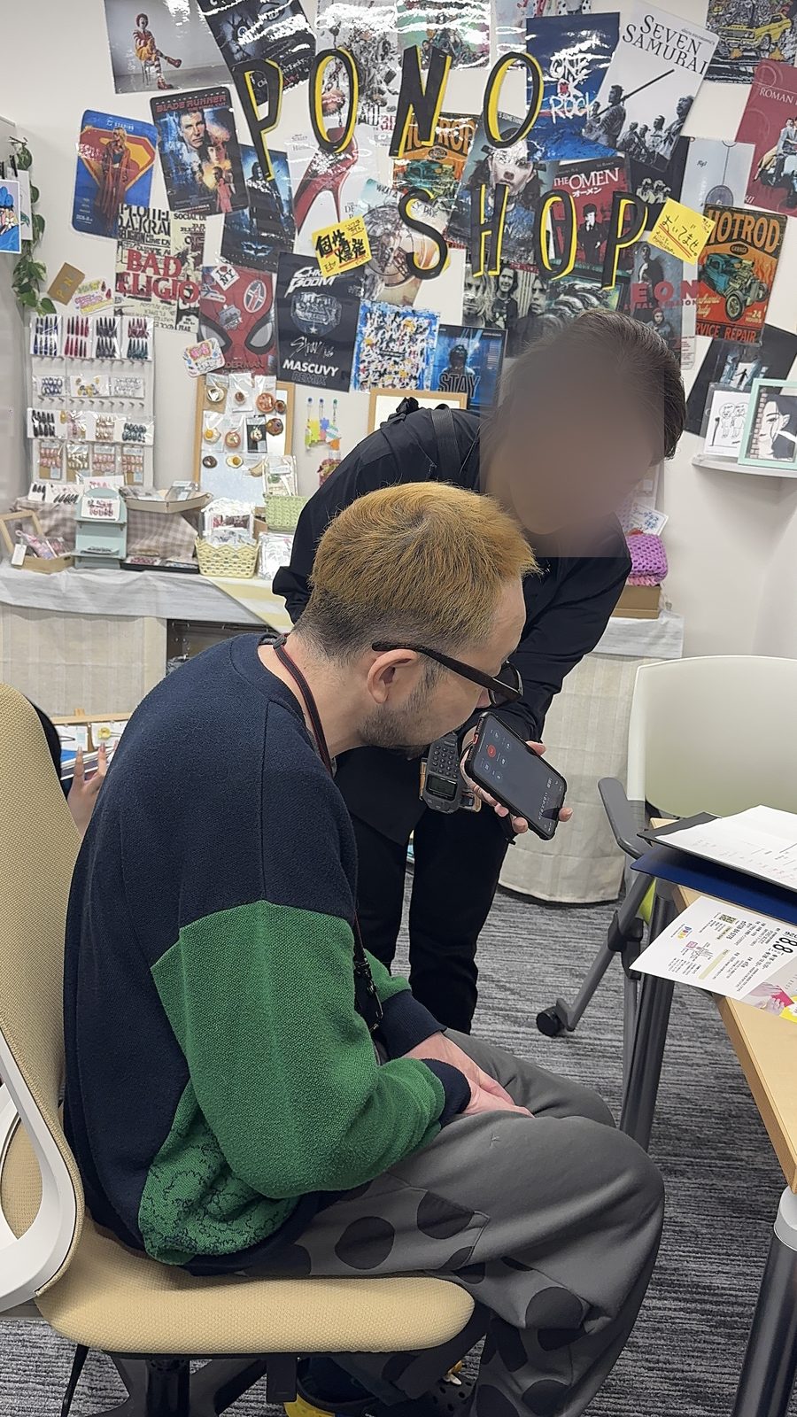 PONO SHOPの装飾がされた壁の前で、ラジオ局の取材者がスマートフォンを向け、PONOの管理者が取材に答えている様子