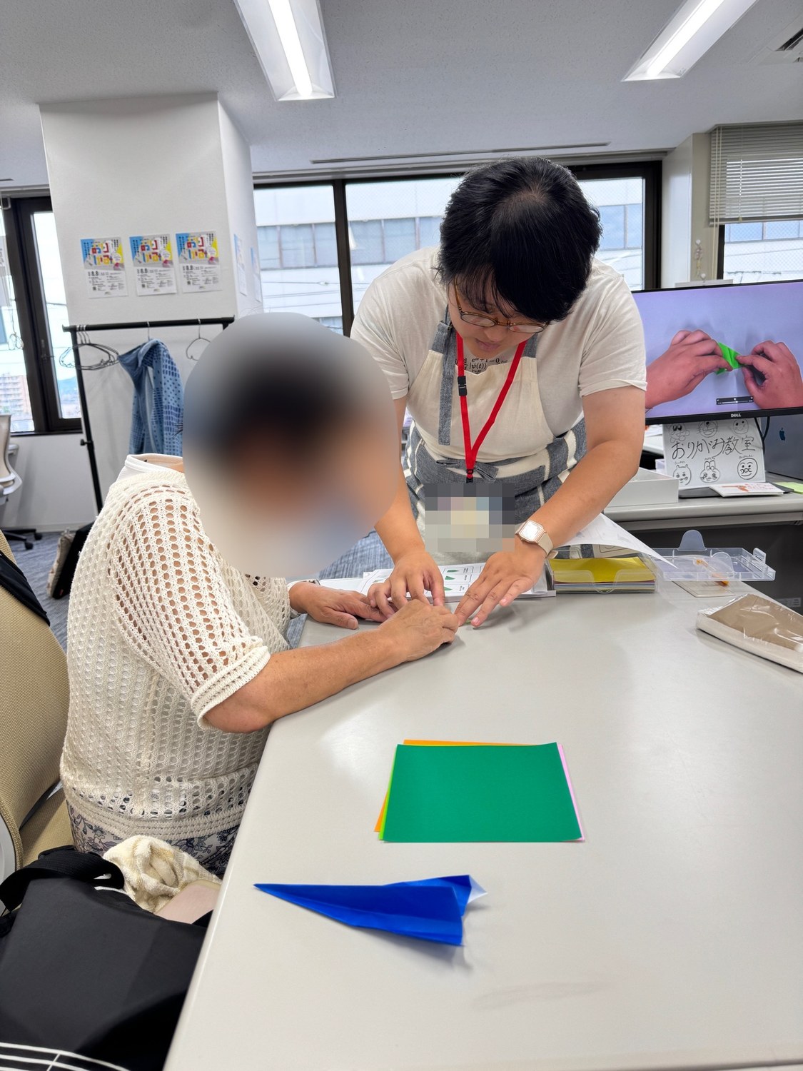 エプロンと名札を身につけた講師が、参加者の隣に立って手元を見ながら折り方を教えている様子。奥のモニターには折り紙を折る手元の動画が映り、その下に手書きの「おりがみ教室」の看板が置かれている